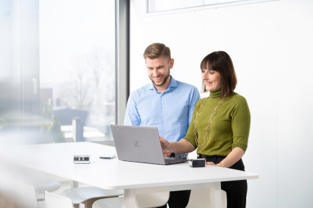 Two employees are standing next to each other in a bright room, looking at a laptop together. On the right is a woman in a green sweater, on the left a young man in a business shirt. Both are smiling.