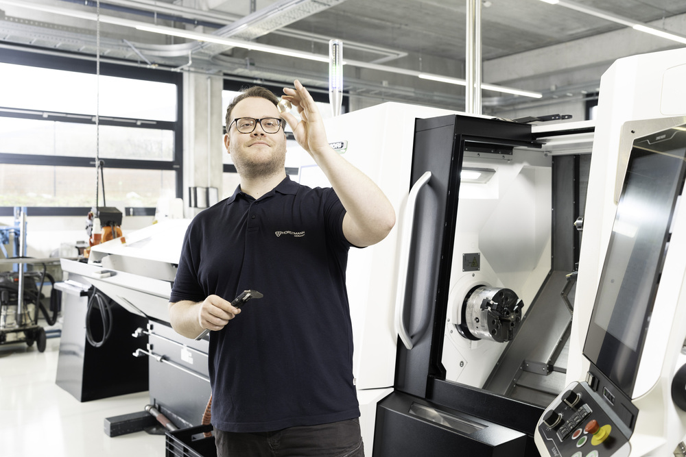 Junger Mitarbeiter aus dem Musterbau hält ein Kupfer-farbiges Kleinteil in die Höhe und prüft es optisch