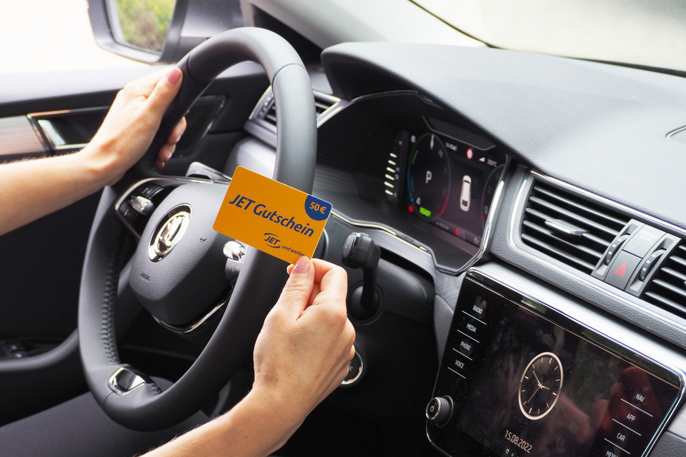 Eine Person sitzt im Auto, hält mit der einen Hand das Lenkrad und in der anderen Hand zeigt Sie eine Tankstellen-Gutscheinkarte in Höhe von 50 Euro