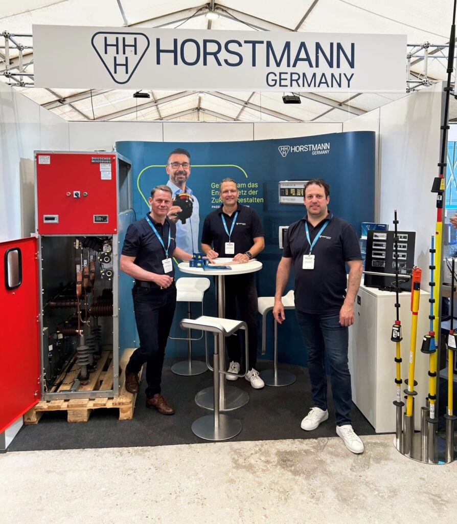 Drei Männer in dunkelblauer Kleidung vor einem mobilen Horstmann Messestand, links eine Schaltanlage, rechts Produkte aufgestellt