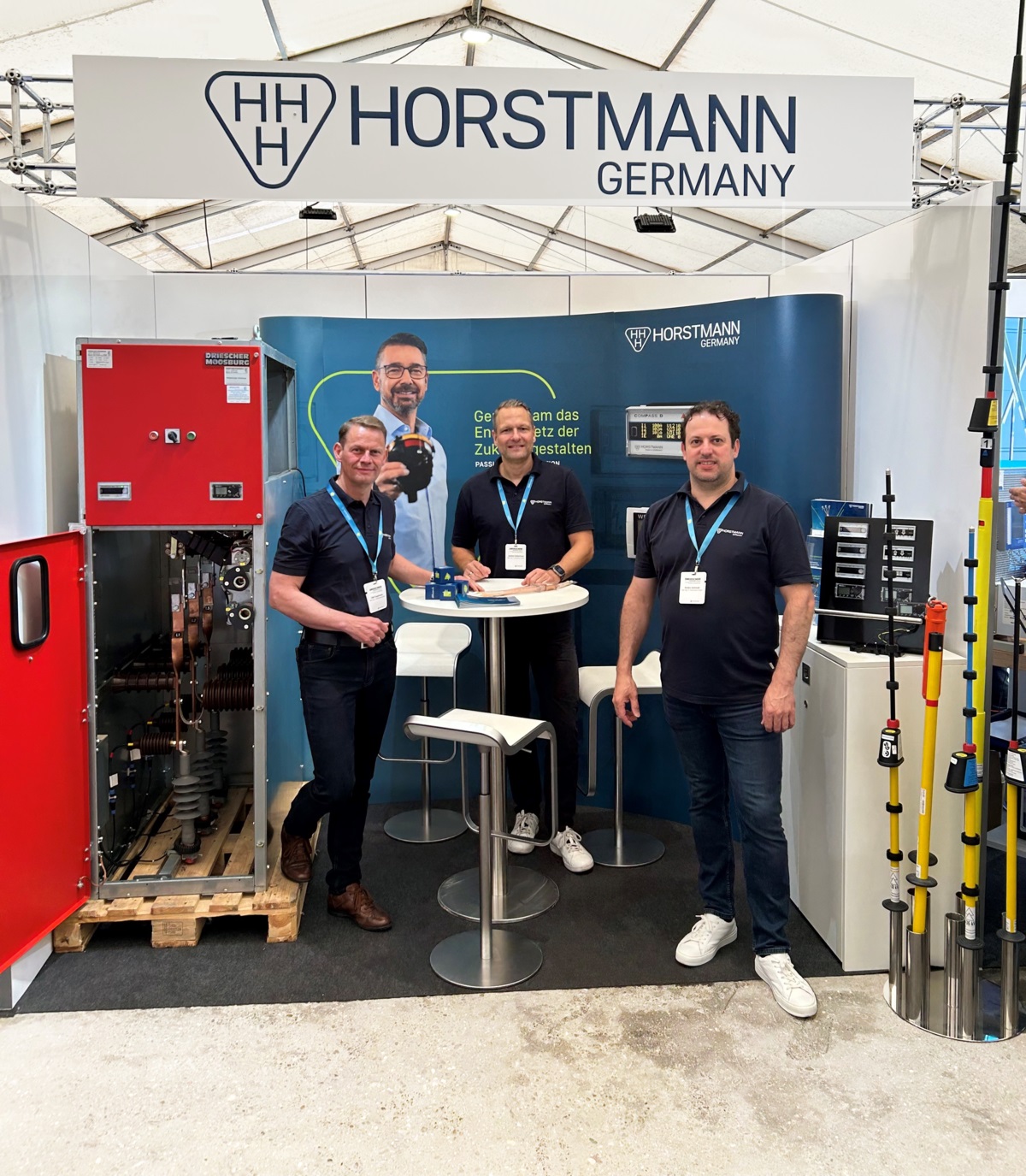 Drei Männer in dunkelblauer Kleidung vor einem mobilen Horstmann Messestand, links eine Schaltanlage, rechts Produkte aufgestellt
